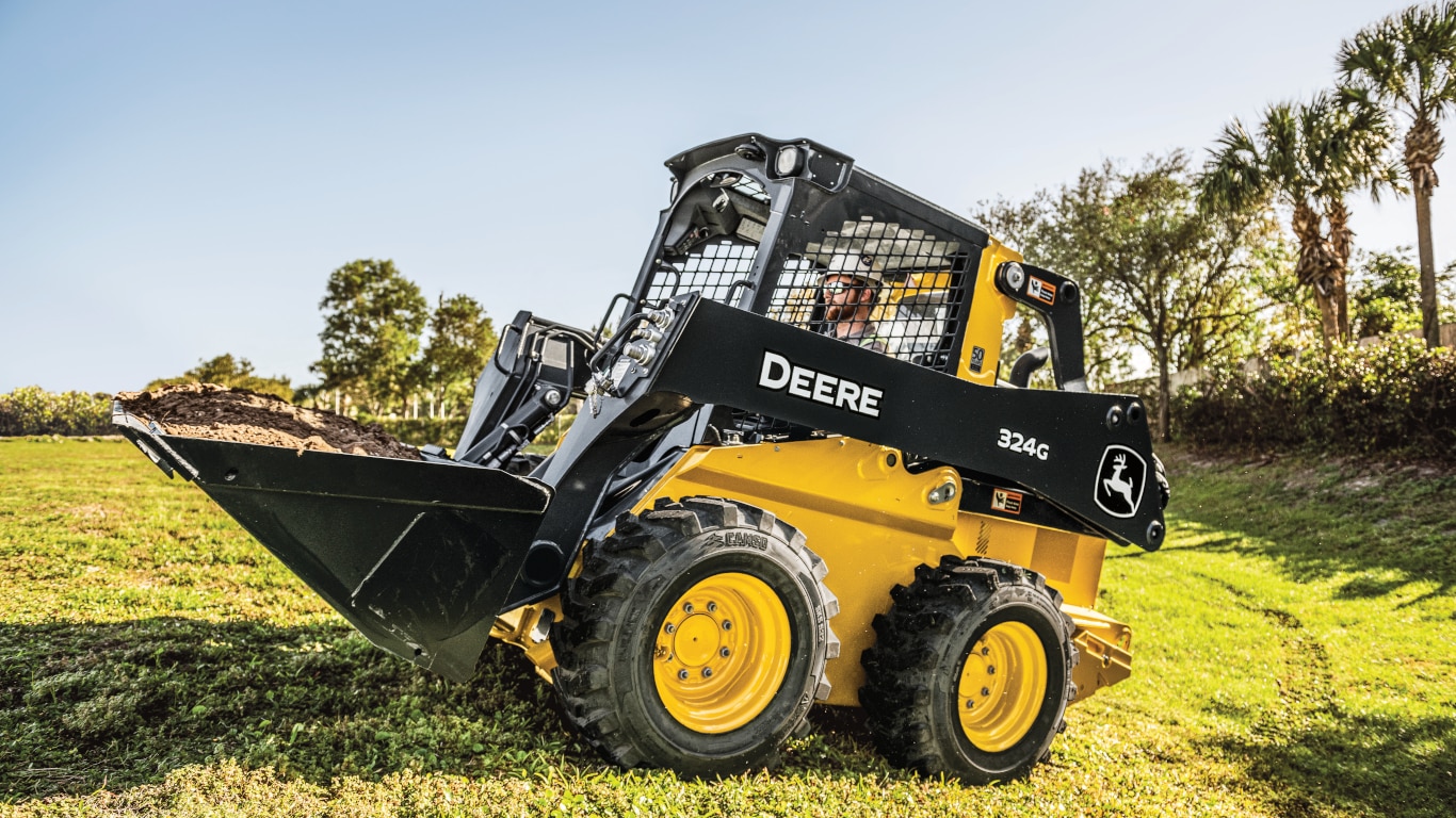 A 324G skid steer transporting dirt on a grassy field with trees in the background.